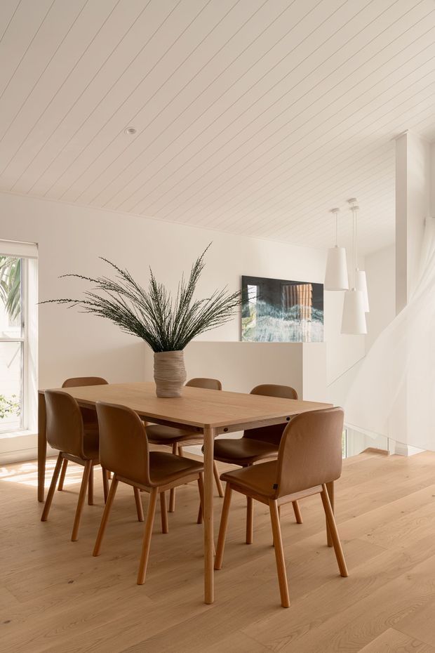 Bright, warm-toned dining area with a large light timber dining table and six brown leather-upholstered dining chairs. The room features pale timber flooring, a white painted ceiling with narrow linear boards, white walls, a stair opening with a sculptural white balustrade, simple white pendant lights, and a large abstract artwork. Natural light enters through full-height glazing at the side, creating a calm, refined contemporary interior.
