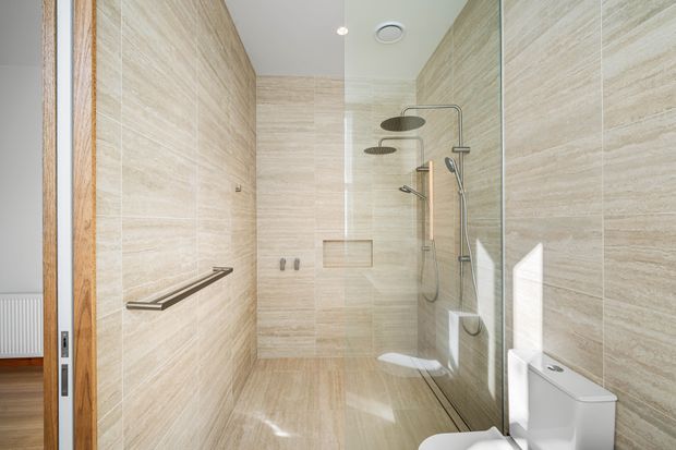 Bright contemporary bathroom with a fully tiled walk-in shower, frameless glass screen, dual overhead rain showerheads, handheld shower, recessed niche, wall-mounted controls, toilet, and timber-framed doorway. The bathroom uses warm beige stone-look tiles throughout with brushed metal fixtures and abundant natural light.
