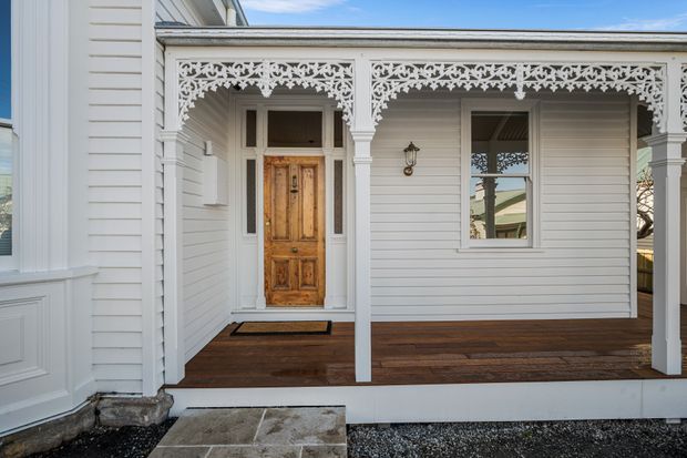 Front entrance porch of a heritage-style weatherboard home, featuring ornate white lacework fretwork, turned porch posts, a richly timber-panelled front door, white horizontal cladding, dark timber decking, and a stone entry step.
