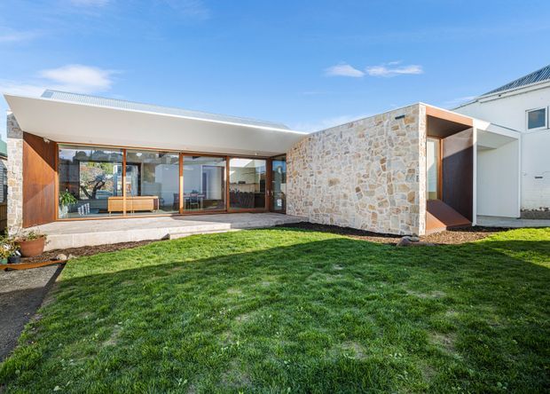 Exterior view of a low, single-storey modern home arranged around a grassy front courtyard. The building combines a broad shallow roof with extensive floor-to-ceiling glazing, warm timber-framed doors and screens, and a prominent irregular natural-stone wall. A recessed entry is visible beside a white rendered wing, with a covered outdoor terrace along the glazed living area.