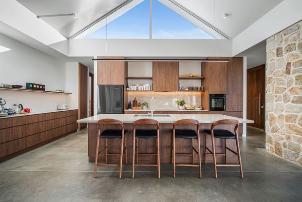 A spacious contemporary kitchen with a large central island and four timber bar stools. The room features dark timber cabinetry, a pale stone benchtop and splashback, integrated appliances, polished concrete flooring, exposed natural stone walling, and a dramatic pitched ceiling with high triangular glazing.