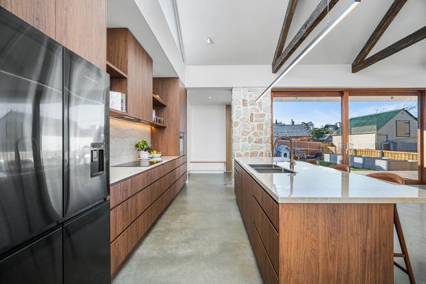 Bright contemporary kitchen with extensive warm timber cabinetry, a large central island with a white stone benchtop, black appliances, polished concrete flooring, exposed timber ceiling beams, and a substantial natural-stone feature wall. Large timber-framed windows open to an urban residential outlook and bring in abundant daylight.