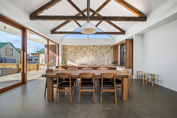 A bright, open-plan dining room with a long solid timber dining table and matching wooden chairs, positioned beneath a high vaulted ceiling with exposed dark timber trusses. Full-height timber-framed glazing opens toward an outdoor area, while a rugged natural-stone feature wall anchors the far end of the room. The space combines polished concrete flooring, white walls, warm timber, and a simple contemporary pendant light.