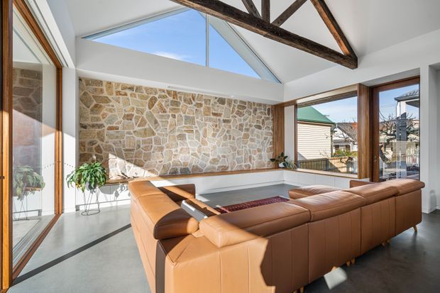 Bright, spacious living room with a large caramel leather sectional arranged around a low central table. The room features a dramatic vaulted ceiling with exposed dark timber beams, extensive glazing, a high clerestory/skylight, polished concrete flooring, and a prominent irregular natural-stone feature wall. Indoor plants and views to neighbouring buildings are visible through the windows.