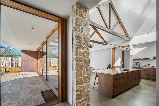 Bright contemporary kitchen and dining area in a renovated heritage-style structure, featuring a large dark timber island, white cabinetry, polished concrete flooring, exposed weathered timber roof trusses, and a substantial exposed stone wall. Full-height timber-framed glazing opens toward a tiled courtyard and allows the original structural elements to remain prominent.