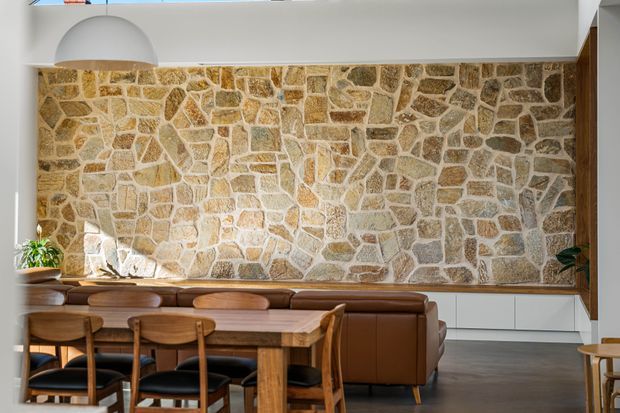 Warm contemporary dining and living area featuring a large irregular natural-stone feature wall, a timber dining table with matching chairs, and a brown leather banquette or sectional sofa. White built-in cabinetry runs along the base of the stone wall, while a sculptural white pendant light and indoor greenery add contrast. The space is well lit with a refined rustic character.