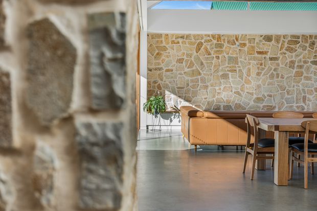 A contemporary open-plan living and dining area viewed beside a rustic stone wall. The space features a polished concrete floor, warm tan leather sofa, timber dining table and chairs, indoor greenery, and a high glazed clerestory/skylight that brings in strong natural light. A distinctive irregular stone feature wall contrasts with the clean modern ceiling and glazing.