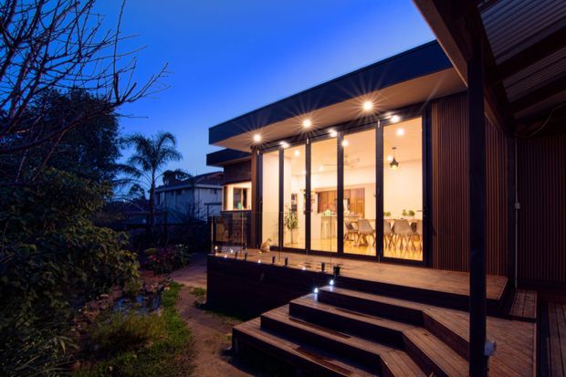 Twilight exterior view of a contemporary single-storey home extension with extensive floor-to-ceiling glazing, a deep flat roof overhang, dark vertical cladding, and a timber deck with broad steps. Warm interior lighting reveals a dining and kitchen area through the glass, while landscaped planting surrounds the structure.