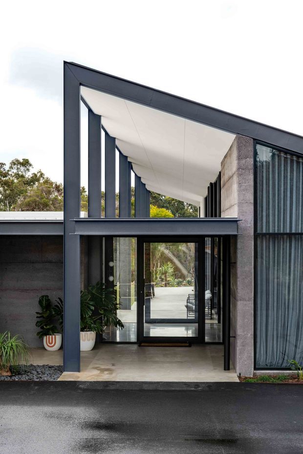 A contemporary residential entrance viewed from outside, framed by a bold dark steel or aluminium portal structure. The entry features floor-to-ceiling glazing, a recessed covered walkway, slender vertical columns, a pale concrete or plaster ceiling, and a textured exposed concrete wall. Dark-framed glass doors open toward a landscaped interior courtyard, with potted plants beside the entrance. The facade has a strong geometric, minimalist composition and a flat roofline.
