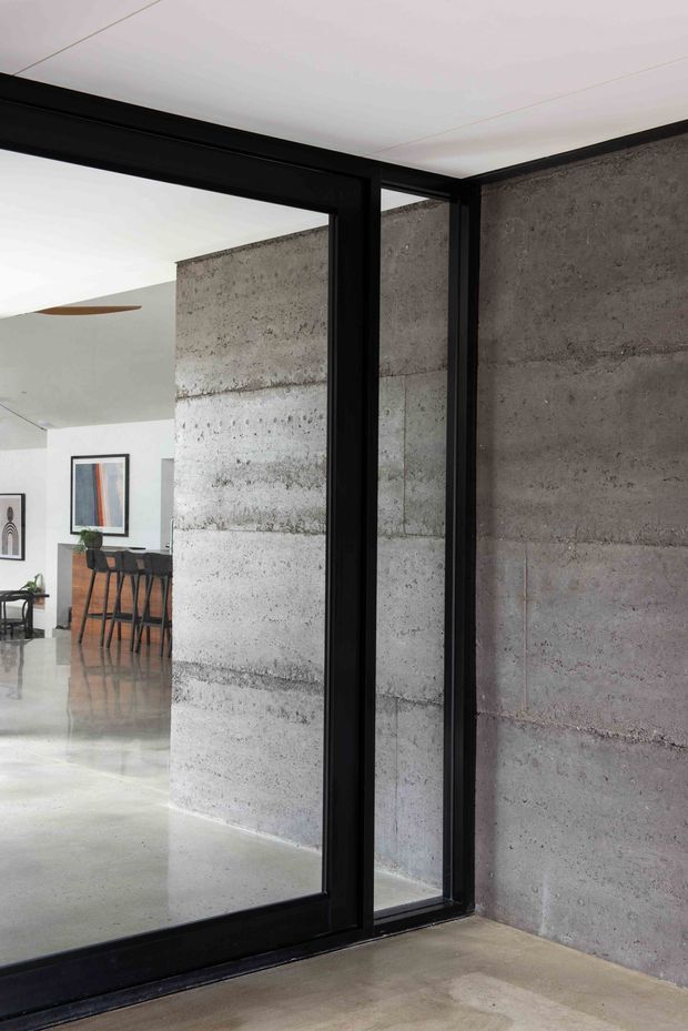Interior architectural view featuring a black-framed glazed partition or sliding door beside exposed grey concrete block walls. Through the opening is a bright open-plan dining and kitchen area with a bar counter, stools, framed artwork, polished concrete flooring, and a minimal white ceiling.