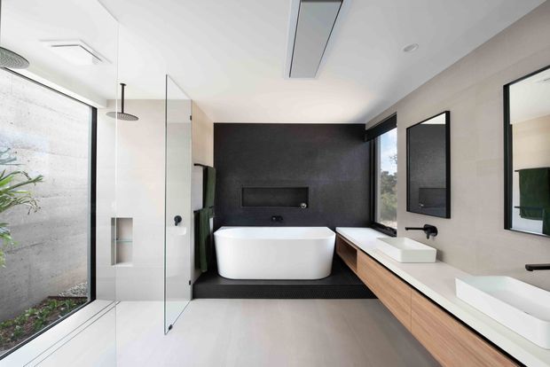 Contemporary spacious bathroom featuring a freestanding white bathtub against a dark textured feature wall, a large double vanity with light timber cabinetry and white basins, walk-in glass shower enclosure, black tapware and fixtures, full-height glazing, and minimalist recessed lighting.