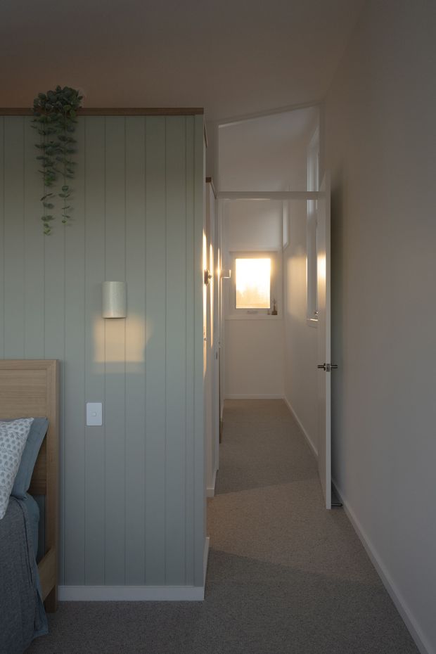 A softly lit bedroom corner opening onto a narrow carpeted hallway. The bedroom features muted sage-green vertical wall panelling, a timber bed frame with blue-grey bedding, a wall-mounted cylindrical light, and trailing greenery. White doors and trim lead toward a bright window at the end of the hall.