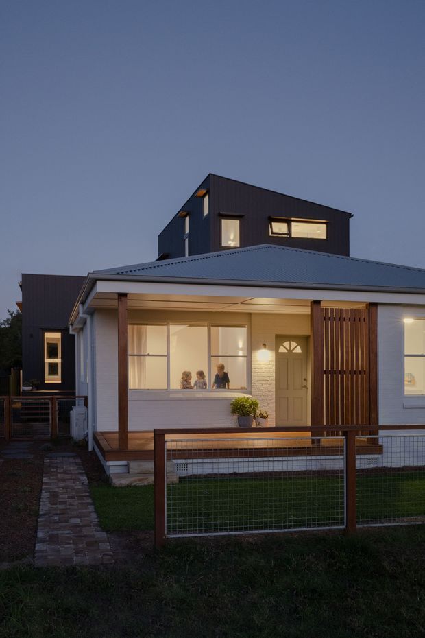 Twilight exterior view of a renovated or extended residential house. The single-storey front portion has a covered veranda, pale painted cladding, a corrugated metal roof, large glazed windows, a timber front door, and a vertical timber privacy screen. A taller dark-clad upper volume rises behind it, creating a contemporary addition. A low timber-and-wire fence and paved path lead toward the entry, with warm interior lighting visible through the windows.