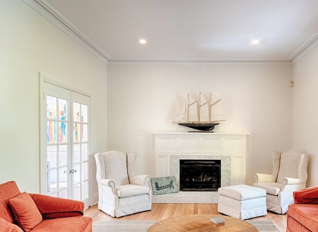 Bright, traditionally styled living room with a white painted fireplace surround and black firebox as the focal point. The room has pale walls with ornate crown moulding, French doors with divided glazing, two cream upholstered armchairs and ottomans, red-orange upholstered sofas, and a warm herringbone timber floor. A model sailing ship sits on the mantel.