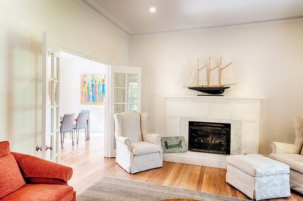 Bright traditional living room with a white paneled fireplace mantel and black firebox as the focal point. The room includes a red upholstered sofa, light upholstered armchair and ottoman, a pale sofa at right, a neutral area rug, and warm timber flooring. French doors open toward a dining area, while a sailing-ship model decorates the mantel. Decorative ceiling cornices and recessed lighting contribute to the classic interior character.