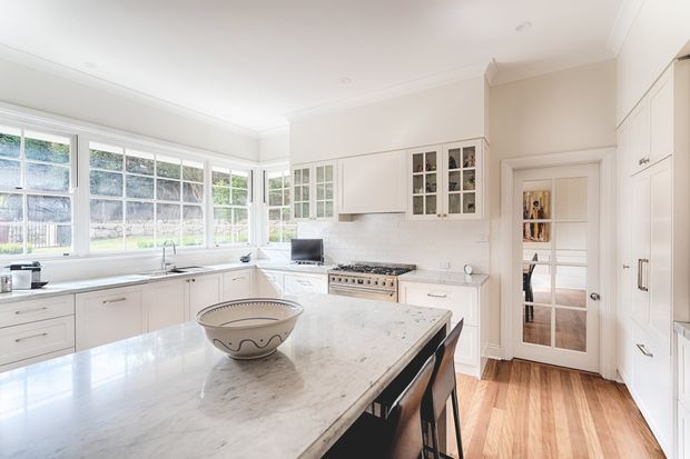 Bright, spacious traditional kitchen with extensive white cabinetry, a large marble island, stainless-steel range, glass-front upper cabinets, and a wall of multi-pane windows overlooking greenery. Warm timber flooring and dark island stools provide contrast, while a glazed interior door leads to a pantry or adjoining area.