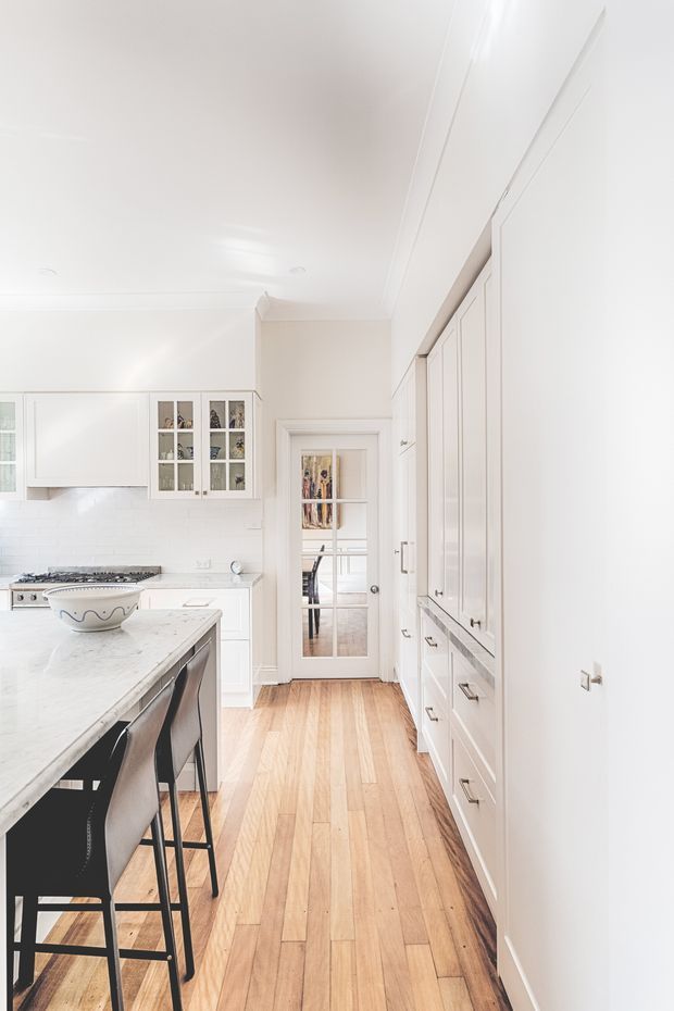 Bright, narrow white kitchen with extensive built-in shaker-style cabinetry, a large pale stone island with dark bar stools, white tiled backsplash, freestanding or integrated cooking area, and warm timber plank flooring. A glazed internal door leads to an adjacent room, while glass-front upper cabinets display crockery or decor. The space has a clean, refined and airy traditional-contemporary character.