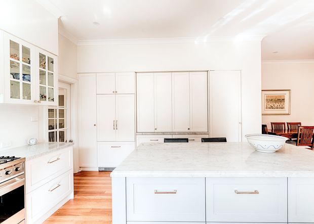 Bright, spacious white kitchen with extensive built-in cabinetry, a large central island with a pale stone benchtop, glass-front display cabinets, integrated appliances, and warm timber flooring. A dining area is visible beyond the kitchen opening.