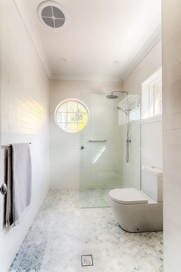 Bright, narrow contemporary bathroom with white horizontal wall tiling, pale hexagonal floor tiles, a frameless glass walk-in shower, wall-mounted rain shower, freestanding-style toilet, chrome towel rail, and a round window that brings in natural light. The space has a clean, light and airy aesthetic.