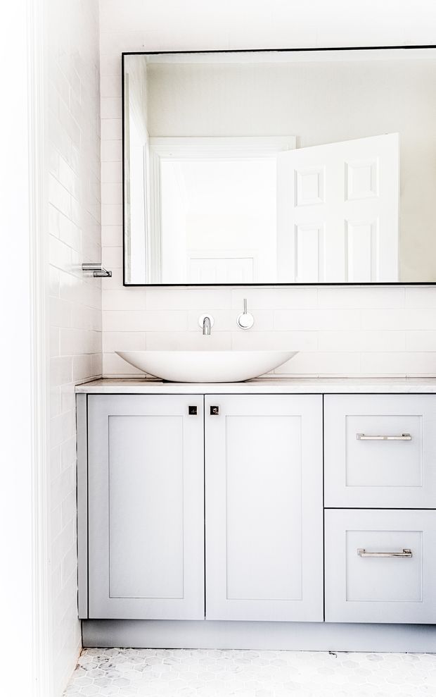 Bright, compact bathroom vanity with a white vessel basin, wall-mounted chrome tapware, white horizontal wall tiles, framed rectangular mirror, pale panelled cabinetry and small hexagonal floor tiles. The mirror reflects a white interior door. The overall look is clean, restrained and contemporary with subtle traditional detailing in the vanity doors.