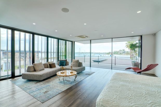 Bright contemporary living room with expansive floor-to-ceiling glazing overlooking a waterfront setting. The room features a large sectional sofa, sculptural lounge chair, round glass coffee table, patterned area rug, and a partially visible bed or daybed in the foreground. Dark timber flooring, white walls and ceiling, and slim dark window frames create a clean, airy interior.