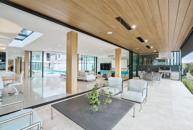 Bright open-plan contemporary interior combining a kitchen, dining area and living room. The space features extensive floor-to-ceiling glazing, black-framed sliding doors, a warm timber-lined ceiling, timber structural columns, polished pale flooring, minimalist furnishings, and a sleek island kitchen with integrated appliances. A pool and exterior views are visible beyond the glazing.