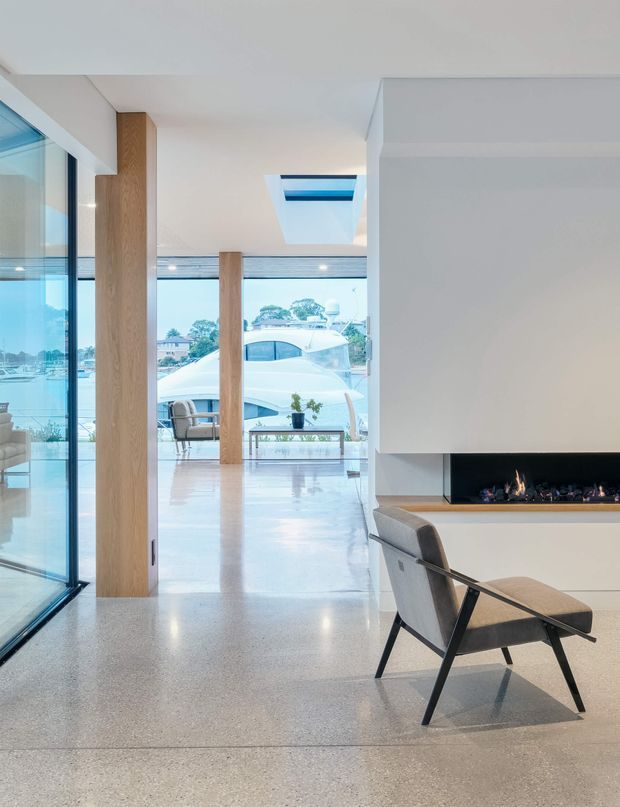Bright, refined contemporary living space with polished concrete or terrazzo-like flooring, extensive floor-to-ceiling glazing, pale timber columns, a large white fireplace volume with a linear built-in fire, and a skylight. A modern lounge chair sits in the foreground, while a waterfront or marina-like view is visible beyond the glazing.