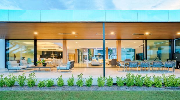 Wide exterior view of a contemporary single-storey residential pavilion with extensive floor-to-ceiling glazing opening onto a landscaped lawn. The home features a broad flat roof with a warm timber-lined soffit, slender dark window and structural frames, and an open-plan interior visible through the glazing, including living and dining areas. Low clipped planting forms a neat foreground border.