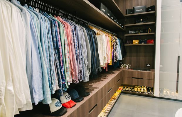 A well-organised walk-in wardrobe with extensive open hanging rails displaying shirts and clothing, dark wood-look shelving and cabinetry, integrated drawers, and dedicated shoe storage. The space has a contemporary, premium custom-joinery feel with a neutral palette and efficient built-in storage.