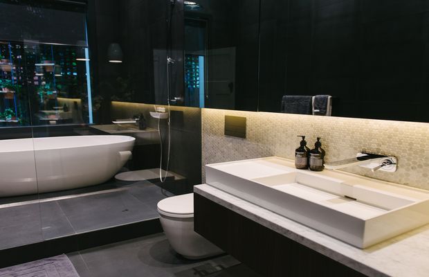 A dark, sophisticated contemporary bathroom featuring a freestanding white bathtub, wall-mounted shower, toilet, and a large double-sink vanity. The room has charcoal-grey floor and wall surfaces, extensive mirror panels, a pale textured mosaic-tile splashback, and a dark timber vanity with a light stone benchtop. Warm concealed lighting illuminates the vanity wall.
