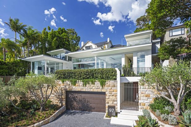 Exterior street-level view of a contemporary multi-level residence with extensive floor-to-ceiling glazing, flat roof planes, white rendered walls, and landscaped grounds. A stone retaining wall, integrated garage, pedestrian entry gate, balconies, and mature tropical planting are visible under a bright blue sky.