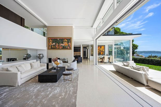 A bright, spacious contemporary living room with a double-height void and expansive floor-to-ceiling glazing opening toward a waterfront outlook. The room features white sofas and armchairs, a dark rectangular coffee table, light neutral flooring, a large grey area rug, and a framed artwork. A second seating area is visible through the glazing, along with landscaped grounds and water beyond.