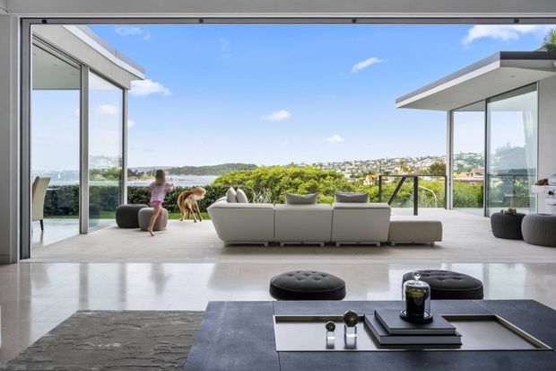 Bright contemporary open-plan living room opening onto a generous terrace through fully glazed sliding doors. A large sectional sofa faces a panoramic leafy suburban outlook, while polished pale flooring, a low dark coffee table, and crisp flat-roofed glazing create a refined indoor-outdoor setting.