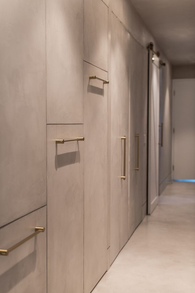 A narrow, softly lit hallway lined with full-height pale timber-toned built-in cabinetry and multiple long brass handles. Smooth light flooring, minimal detailing, and a doorway at the far end create a refined, warm contemporary interior.
