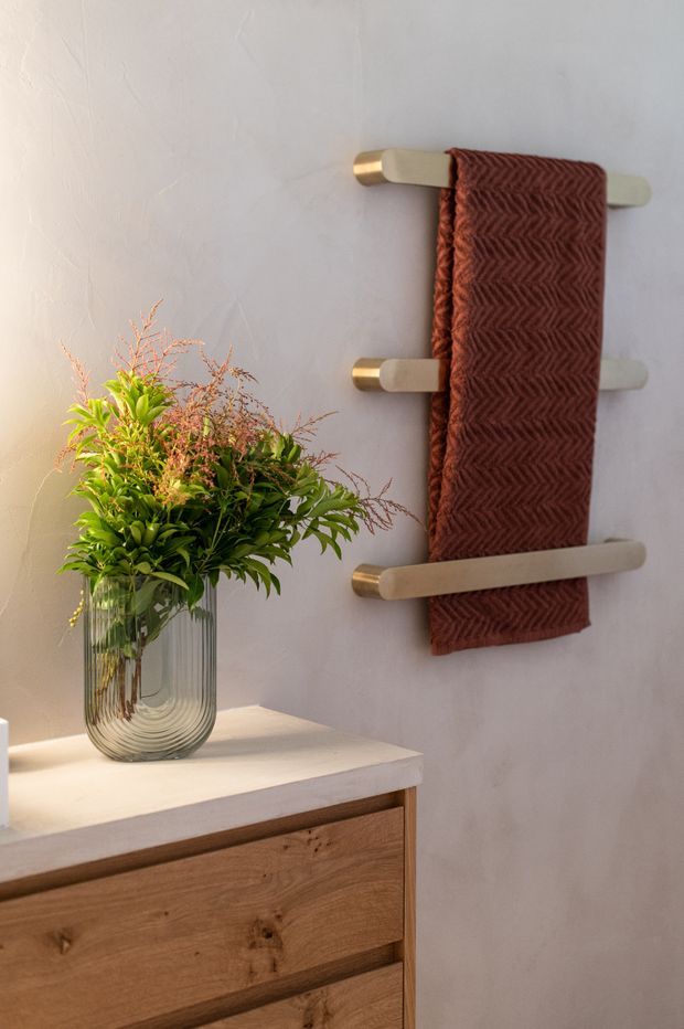 Close-up of a warm, refined bathroom vignette featuring a wall-mounted brushed brass towel rail with a textured rust-brown towel, above a timber vanity with a pale stone benchtop. A clear glass vase of leafy greenery adds a natural accent. The walls are softly textured and light neutral in colour.