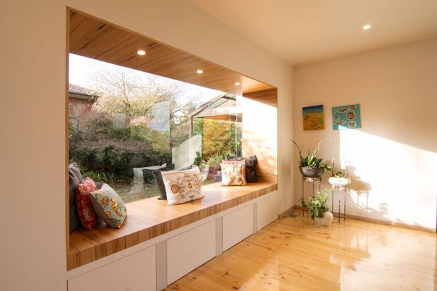 Bright, warm contemporary living space with a large timber-framed picture window and integrated window seat overlooking a planted courtyard. The seat incorporates white built-in storage cabinetry below, while polished timber flooring and a timber-lined reveal add warmth. A small plant display and colourful artwork occupy the otherwise minimal room, with strong afternoon sunlight creating reflections across the floor.