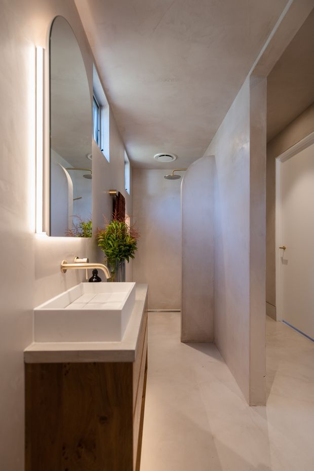 A narrow contemporary bathroom with a sculptural walk-in shower partially screened by curved plaster walls. The room features warm beige plaster walls and floor, a wall-mounted rectangular white basin on a dark timber vanity, a brass tap, arched illuminated mirror, recessed ceiling lighting, and a small high window. A separate doorway is visible at the right.