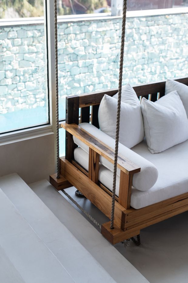 A bright interior seating area featuring a suspended wooden swing sofa with thick rope supports, white upholstered cushions, and large windows. A pale stone or concrete wall and turquoise water are visible outside, creating a clean, airy connection to the outdoors.
