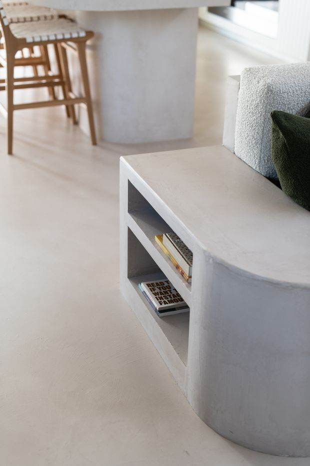 Close-up interior view of a minimalist living area featuring a curved pale concrete or plaster built-in sofa base with open shelving, textured light upholstery, and a dark green cushion. A light-toned kitchen or dining area with timber bar stools is visible in the background.