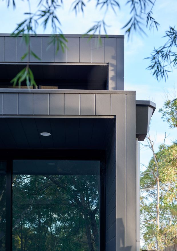 Close-up exterior architectural view of a contemporary dark-clad, multi-level building. Strong horizontal and vertical geometric lines frame deep recessed openings, a projecting balcony or terrace, dark soffits with recessed circular lighting, and large reflective glazing. The facade is set against a leafy treed surroundings and clear sky.
