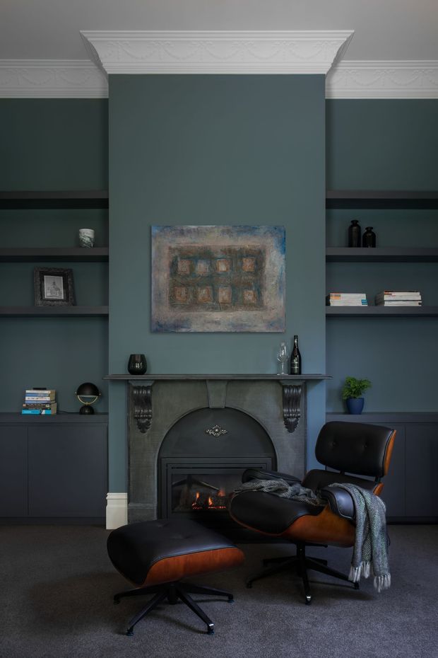 A refined, moody sitting area centred on a dark painted fireplace with built-in shelving on both sides. The deep blue-grey walls and cabinetry are topped with ornate white decorative cornices. A black and warm brown leather lounge chair and ottoman sit on a dark carpet in front of the fireplace, with a muted artwork above and restrained decorative objects and books on the shelves.