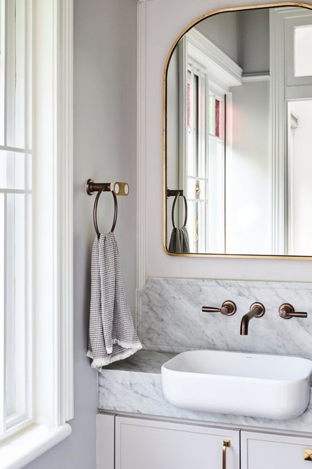 A refined bathroom vanity photographed beside a tall window. The space features a white vessel sink set into a pale grey-white marble countertop and backsplash, bronze wall-mounted tapware, a large rounded-rectangle mirror with a slim brass-toned frame, and light grey cabinetry with brass hardware. Traditional moulded trim and bright natural light create an elegant, classic atmosphere.