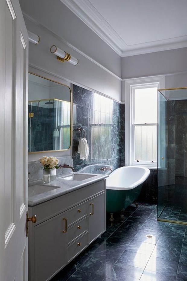 A refined traditional bathroom featuring a grey painted vanity with brass hardware, marble benchtop, inset basin, gold-framed mirror, freestanding bathtub, and glass shower enclosure. Dark veined stone tiles cover the floor and portions of the walls, while white decorative mouldings and a high detailed ceiling add classic character. Natural light enters through the tall window.
