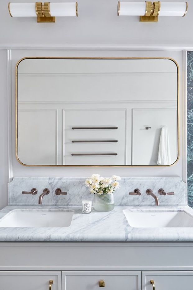 A refined double-sink bathroom vanity in a light grey and white palette. The built-in vanity features a white marble countertop and backsplash, two integrated rectangular basins, brushed metal wall-mounted faucets, paneled cabinetry with brass knobs, and a large rounded-rectangle mirror with a brass frame. Matching brass-and-white wall sconces sit above the mirror, while a small vase of flowers and hand towel add styling details.