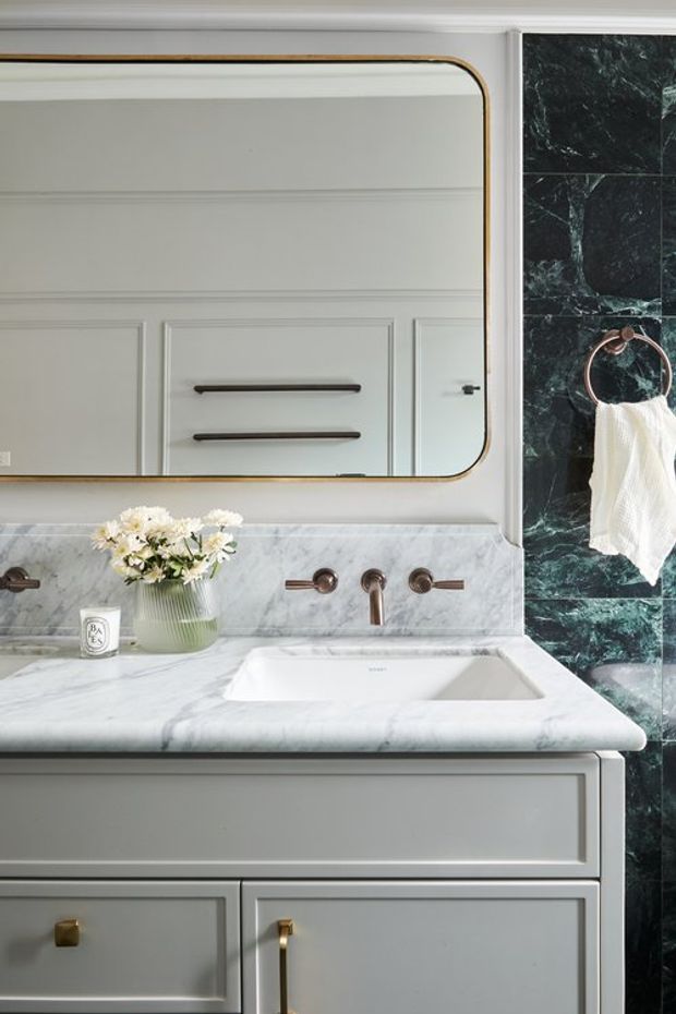 Close-up of an elegant bathroom vanity with pale grey paneled cabinetry, a white-grey marble countertop and integrated rectangular basin, brushed bronze tapware, and a large rounded-corner mirror with a brass-toned frame. Dark green veined marble wall tiles and a matching bronze towel ring provide contrast, while a small flower arrangement and candle add softness.