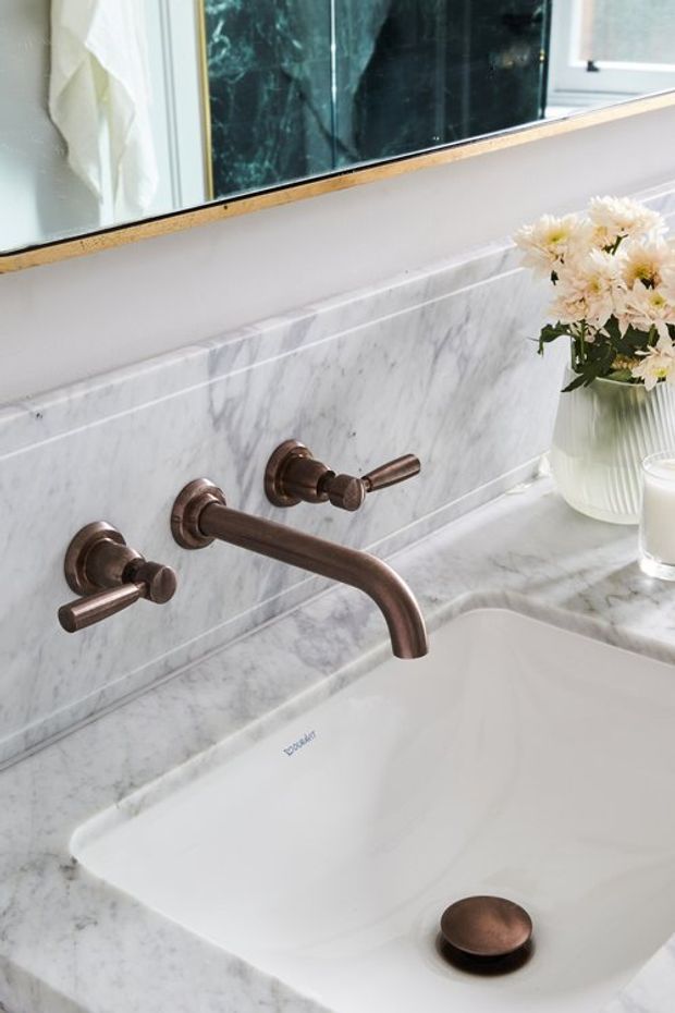 Close-up view of a refined bathroom vanity featuring a white integrated basin, wall-mounted bronze tapware, and a pale veined marble benchtop with matching marble splashback. A large framed mirror and a vase of flowers sit above and beside the basin.