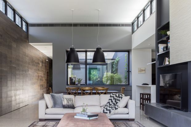 A bright contemporary double-height living room with a large white sectional sofa, timber coffee table, black pendant lights, and a dark built-in media wall. Exposed dark timber wall cladding contrasts with white walls, clerestory glazing, and a large rear window overlooking greenery. A dining area and internal courtyard or garden view are visible beyond the living space.