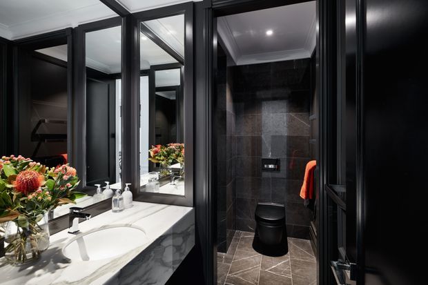 A dramatic, refined bathroom with a double vanity and white marble-effect benchtop, framed mirrors, and black cabinetry and window or door frames. A separate toilet enclosure is finished in dark stone-look wall and floor tiles, with a black toilet and recessed ceiling lighting. A colourful floral arrangement adds a small accent beside the vanity.