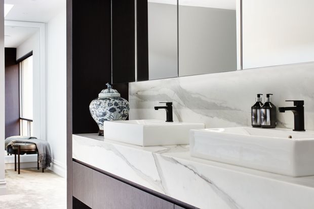 Contemporary bathroom vanity with two white vessel basins, matte black wall-mounted taps, and a dramatic white marble-look stone benchtop and splashback. Mirrored cabinetry spans the upper wall, while dark timber cabinetry and a decorative blue-and-white vase add contrast. A connected bedroom or dressing area is visible through the opening at left.