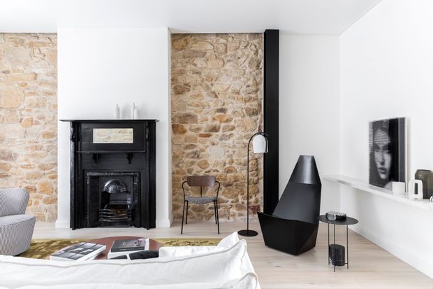 Bright contemporary living room featuring exposed natural stone walls, a restored black fireplace, pale timber flooring, white walls and ceiling, and sculptural black furnishings. A sofa and coffee table occupy the foreground, with a lounge chair, floor lamp, media ledge and framed artwork completing the room.